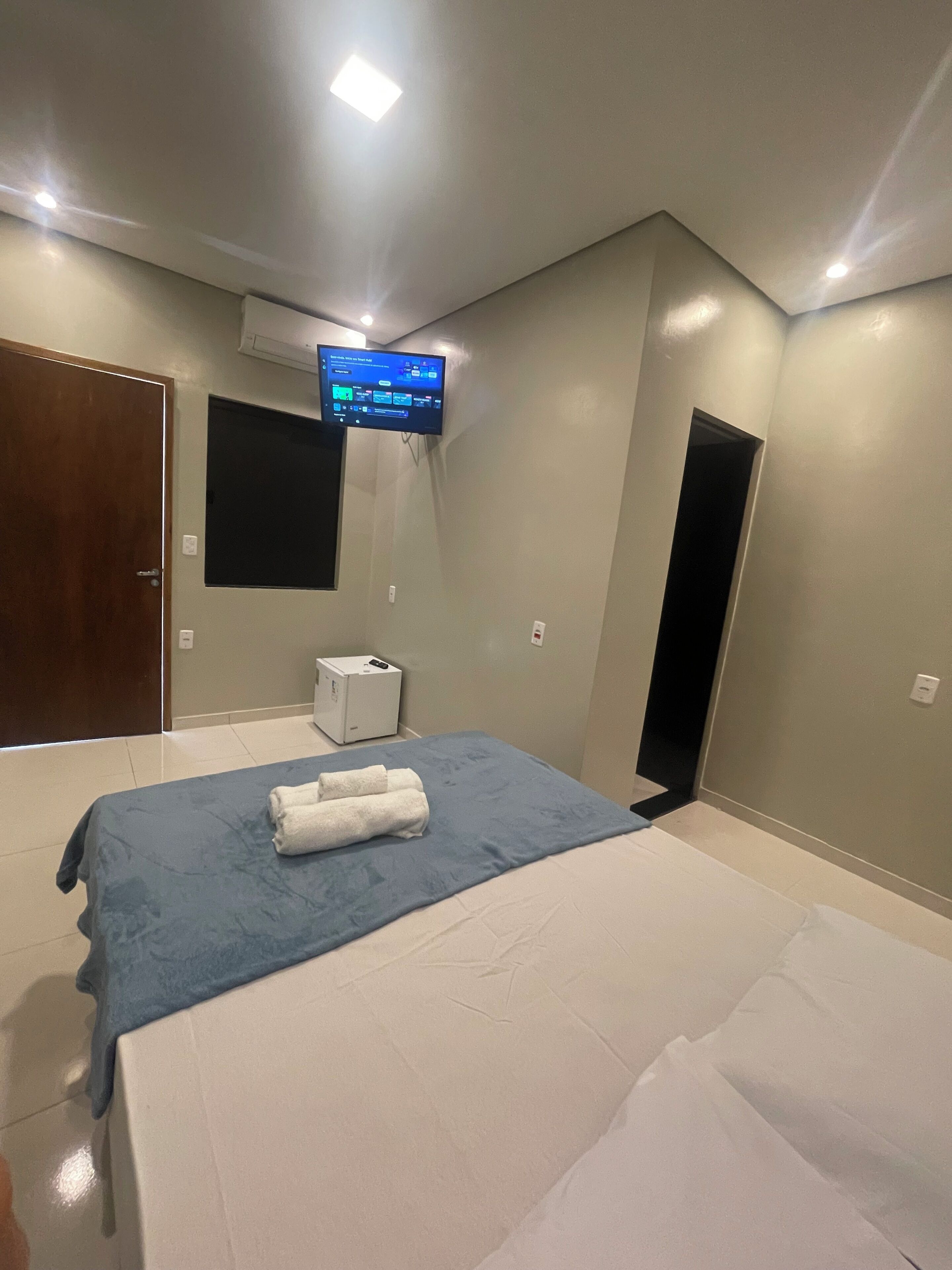 A hotel room with a bed, a television mounted on the wall, a small air conditioning unit, and a door.