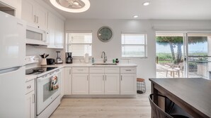 Cocina moderna con armarios blancos, horno empotrado, microondas y fregadero. Hay un reloj en la pared y vistas al exterior a través de la ventana.