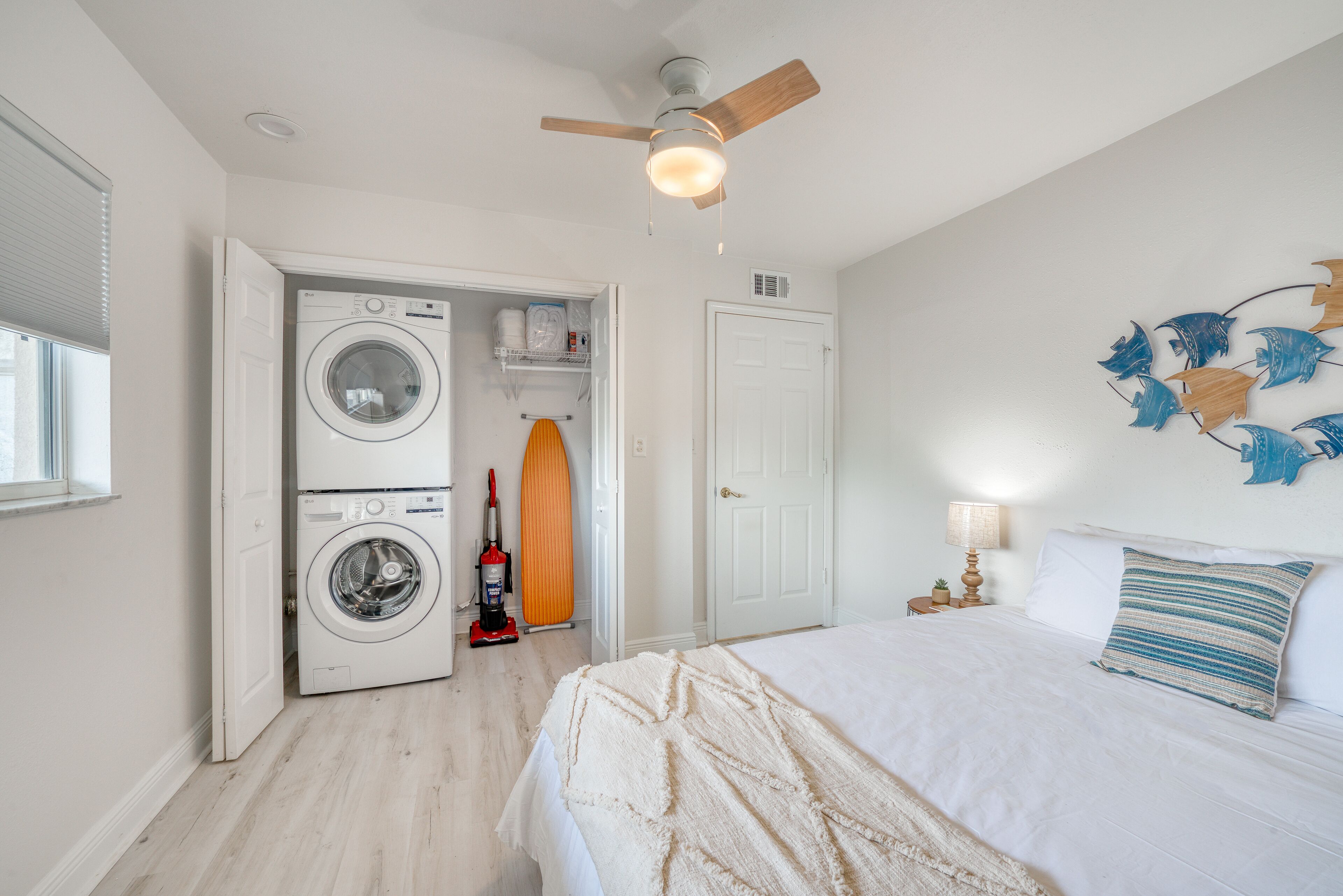 A bedroom with a bed, a ceiling fan, a washing machine, and a vacuum cleaner.