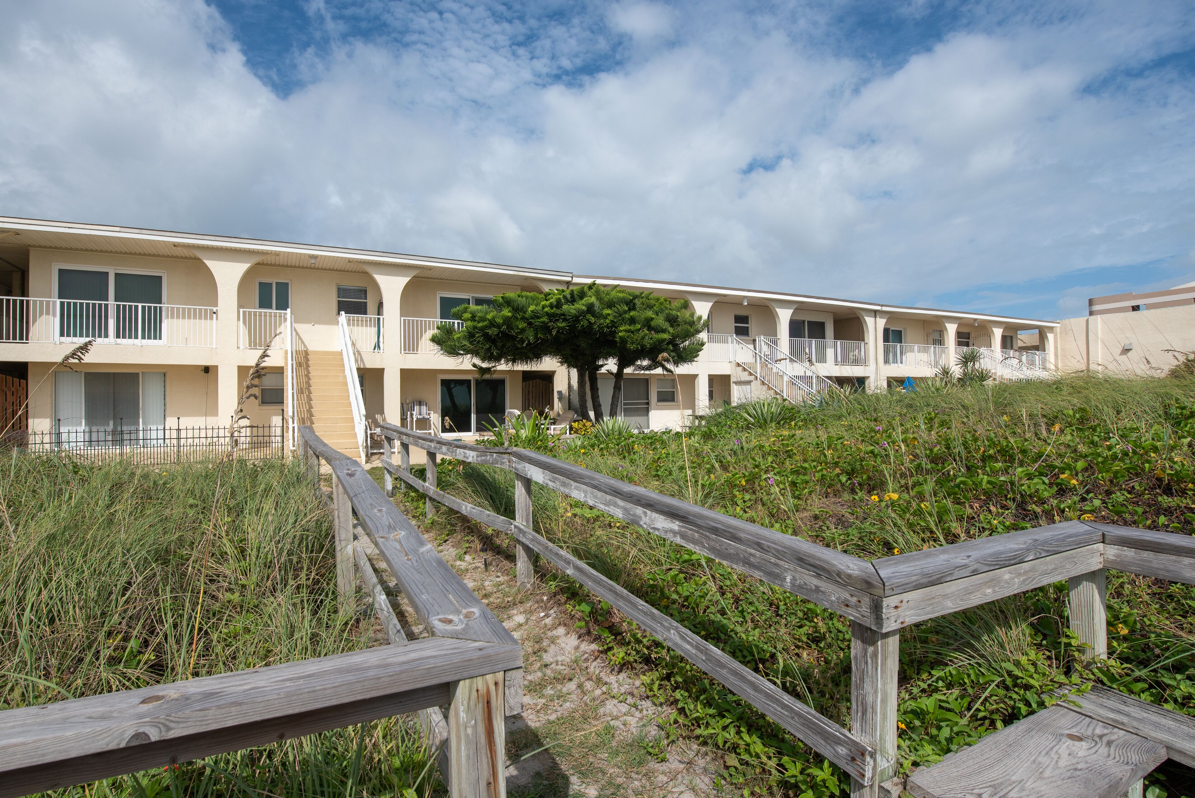A beachfront motel with multiple balconies, a wooden boardwalk, and a large tree in the center.