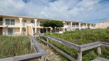 A beachfront motel with multiple balconies, a wooden boardwalk, and a large tree in the center.