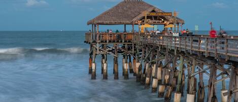 A wooden pier with a thatched-roof structure, likely a restaurant or bar, extending into the ocean.