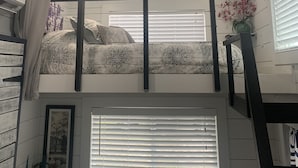 Een kamer met een stapelbed, een raam met zonwering, een plafondventilator en een lijst met een afbeelding aan de muur.