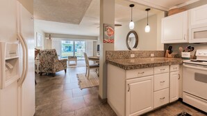 A kitchen with white cabinets, granite countertops, and a built-in oven. There is a dining area with a chair and table, and a view of the outdoors through a glass door. - Off Season - Steep DIscount - Heated Pool (Indialantic)