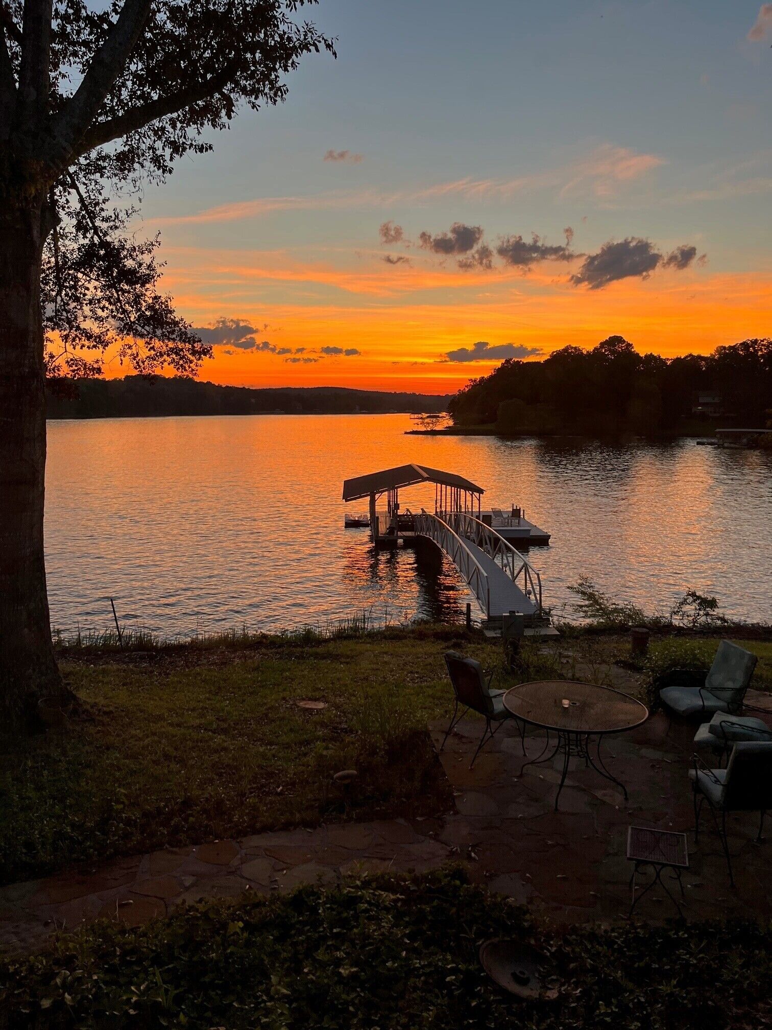 Um alpendre à beira do lago com um cais que se estende sobre a água, rodeado por árvores e uma vista do pôr do sol.