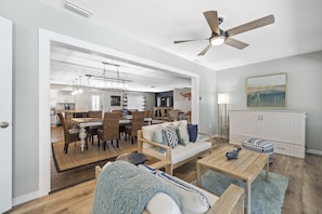 A spacious living room with a wooden floor, a ceiling fan, a dining area with a table and chairs, and a kitchen in the background.