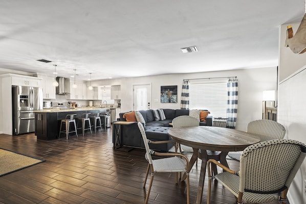 A modern kitchen with stainless steel appliances, a dining area with a wooden table and wicker chairs, and a dark-colored sofa with a striped throw.