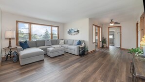 A spacious living room with a large sectional sofa, a small ottoman, and a coffee table. There are large windows providing natural light and a view of trees. A ceiling fan and a wall-mounted TV are visible.