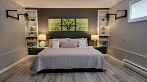 A bedroom with a large bed, two bedside tables with lamps, a wall-mounted shelf with decorative items, and a window with a view of trees.