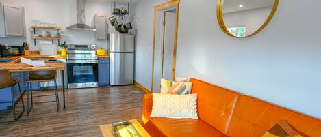 A modern kitchen with stainless steel appliances, a dining area with a wooden table and chairs, and an orange sofa with decorative pillows.
