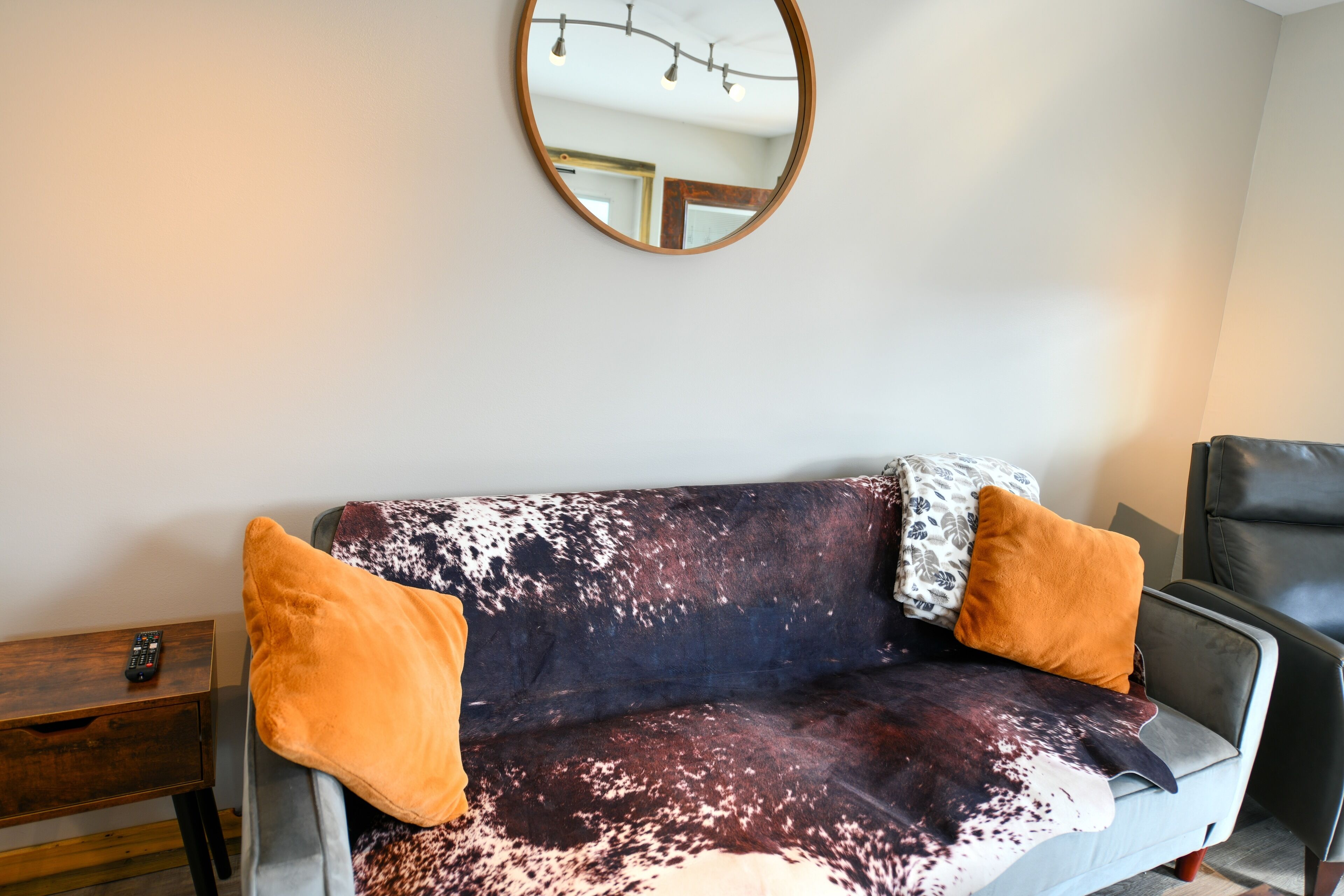 Ein Sofa mit Kuhfellmuster, zwei orangefarbene Kissen und ein runder Spiegel an der Wand.