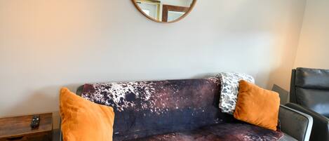 Un sofá con estampado de piel de vaca, dos cojines naranjas y un espejo redondo en la pared.