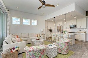 A spacious living room with a sofa, round coffee table, and patterned ottomans. A kitchen with white cabinets and a central island is visible in the background.