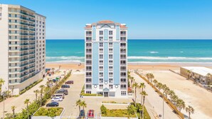 A high-rise hotel with multiple floors and balconies, located adjacent to a beach with a parking lot and palm trees.