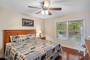 Un dormitorio con cama de madera, ventilador de techo, ventana con persianas y un cuadro en la pared.