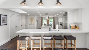 Cocina moderna con una isla central, taburetes de bar, iluminación colgante y armarios blancos.