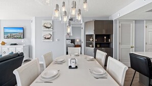A modern dining area with a white table set for four, surrounded by white chairs. There are hanging light fixtures above the table, and a television is mounted on the wall to the left. - Ashworth 309-Oceanfront 4BR Near Main Street, Pool (North Myrtle Beach)