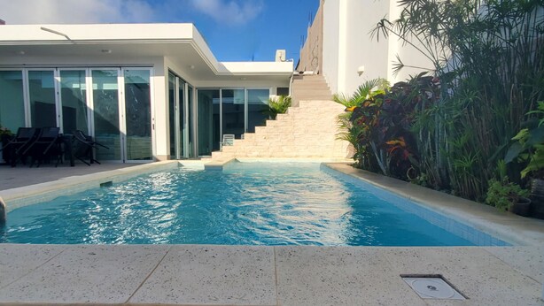 Una piscina moderna al aire libre con superficie de agua azul cristalina, rodeada por un edificio blanco con grandes puertas de vidrio y una escalera que lleva a un nivel superior.