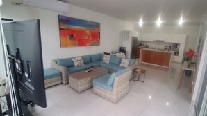 Una sala de estar moderna con un sofá seccional, una mesa de centro y una mesita auxiliar. Se ve una cocina al fondo, con un refrigerador y armarios.