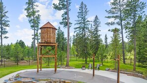 A wooden structure with a conical roof, surrounded by a paved area with stone accents, set in a park with tall trees and a fenced perimeter. - The Fireside-Hot Tub, Firepit, Fireplace (Lead)