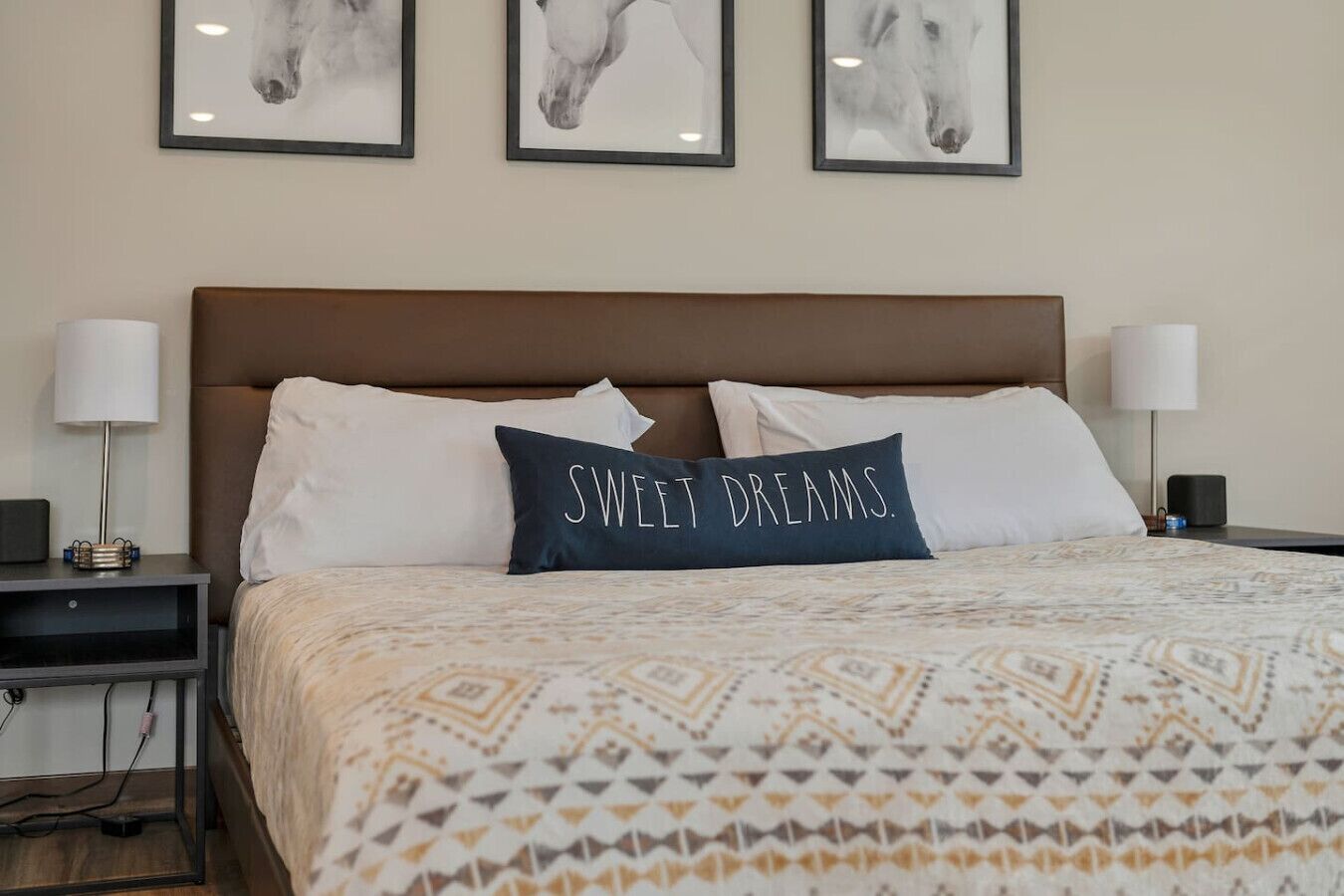 En snyggt bäddad säng med ett mönstrat överkast, två vita kuddar och en mörk kudde med texten ”SWEET DREAMS”. På väggen ovanför sängen hänger tre inramade porträtt av hästar.