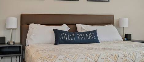 En snyggt bäddad säng med ett mönstrat överkast, två vita kuddar och en mörk kudde med texten ”SWEET DREAMS”. På väggen ovanför sängen hänger tre inramade porträtt av hästar.