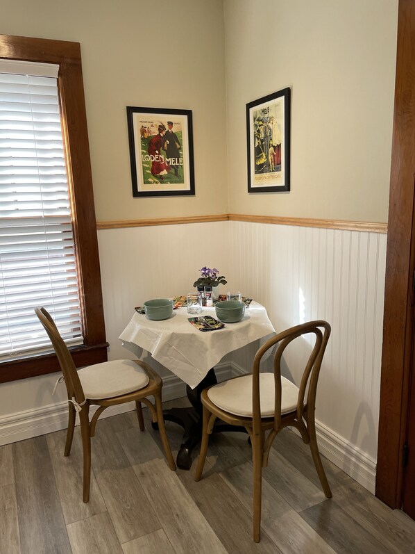 A dining area with a table set for two, two chairs, a window with blinds, and two framed posters on the wall. - Victorian Charm—Studio/Apartment in Delhi Village (Delhi)
