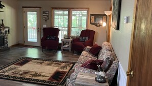 A spacious living room with a fireplace, two red armchairs, a floral sofa, and a large window with a view of greenery. - Relax on the Caddo (Caddo Gap)