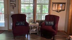 A room with two red armchairs facing a wooden table with books, a window with a view of greenery, and a cabin rules sign on the wall. - Relax on the Caddo (Caddo Gap)