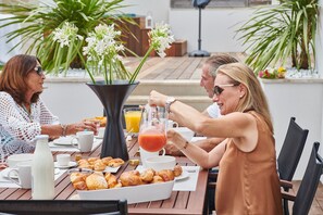 A group of people enjoying breakfast at an outdoor table with croissants, juice, and flowers.