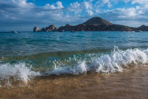 A beach with clear water, waves, and rocky formations in the distance.