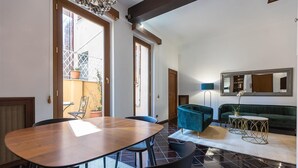 A spacious room with a wooden dining table, black chairs, a teal sofa, and a green armchair. There is a large mirror on the wall and a potted plant on the windowsill.