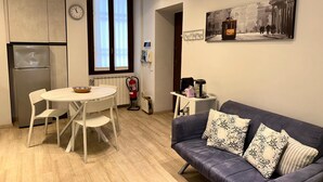 Een moderne keuken met een koelkast, een eettafel met stoelen, een raam met zonwering, een brandblusser, een koffieapparaat en een bank met decoratieve kussens.