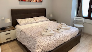 Een net opgemaakt bed met bloemenbedrukking, een nachtkastje met een lamp, en een raam met gordijnen.