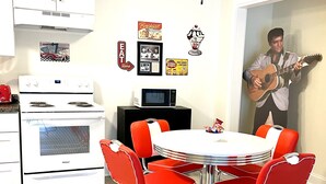 Cucina con piano cottura bianco, forno a microonde e tavolo da pranzo con sedie rosse. Decorazioni a parete tra cui una foto incorniciata e un cartello con la scritta 'Colazione Qui'.
