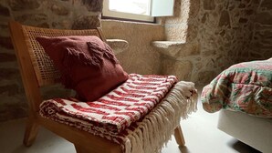 Une pièce chaleureuse avec un mur en pierre, une chaise en bois avec un coussin rouge, une petite fenêtre et un lit recouvert d’une courtepointe à motifs floraux.