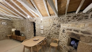 Une pièce chaleureuse avec un mur en pierre, une cheminée, une table à manger en bois avec des chaises, un téléviseur posé sur un meuble en bois et un lustre.