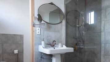 Un baño moderno con un lavamanos pedestal, un espejo redondo, cabina de ducha de vidrio y una cesta de mimbre en el suelo.