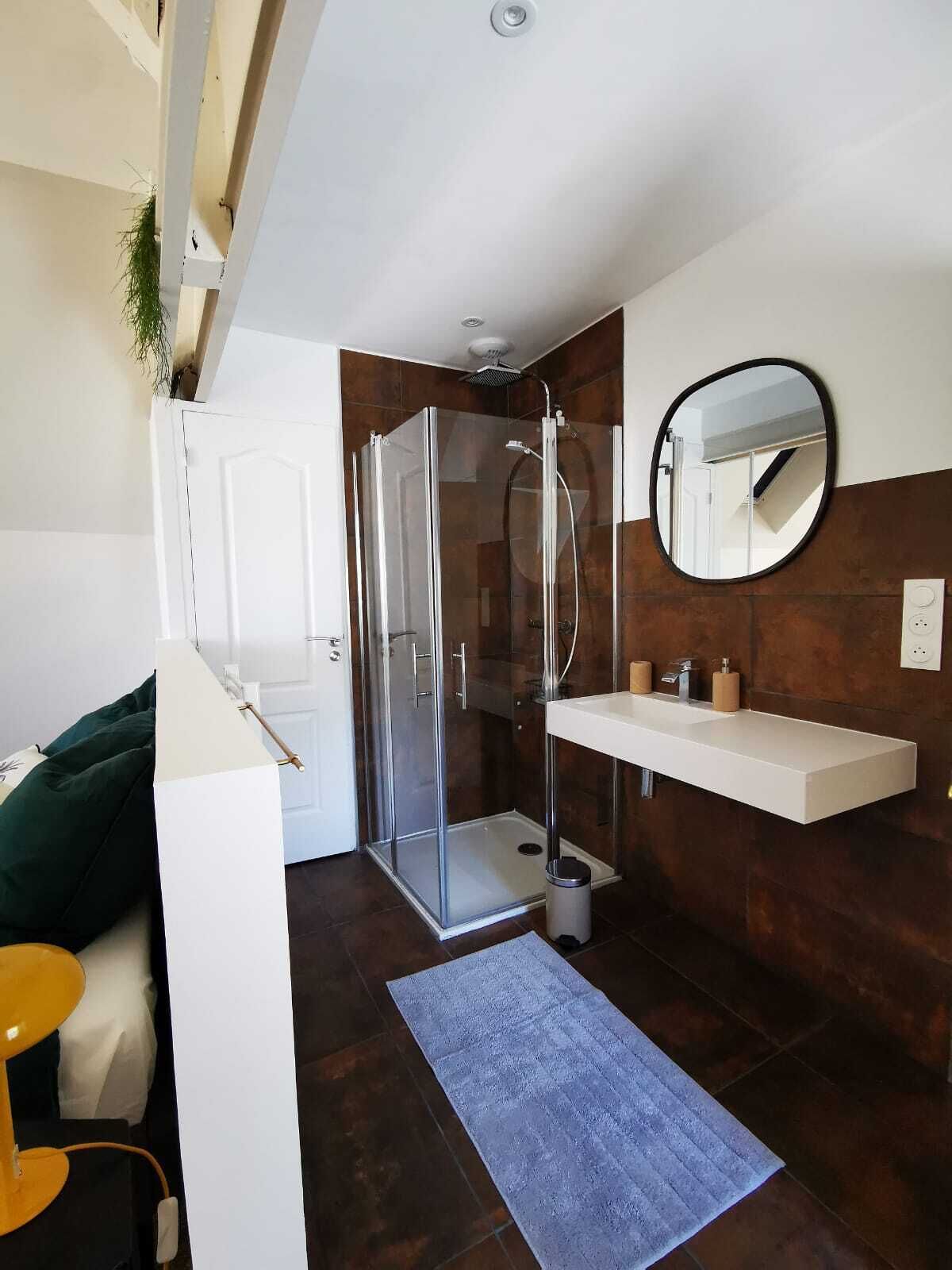 A modern bathroom with a glass-enclosed shower, a white sink with a mirror above it, and a dark brown tiled floor.