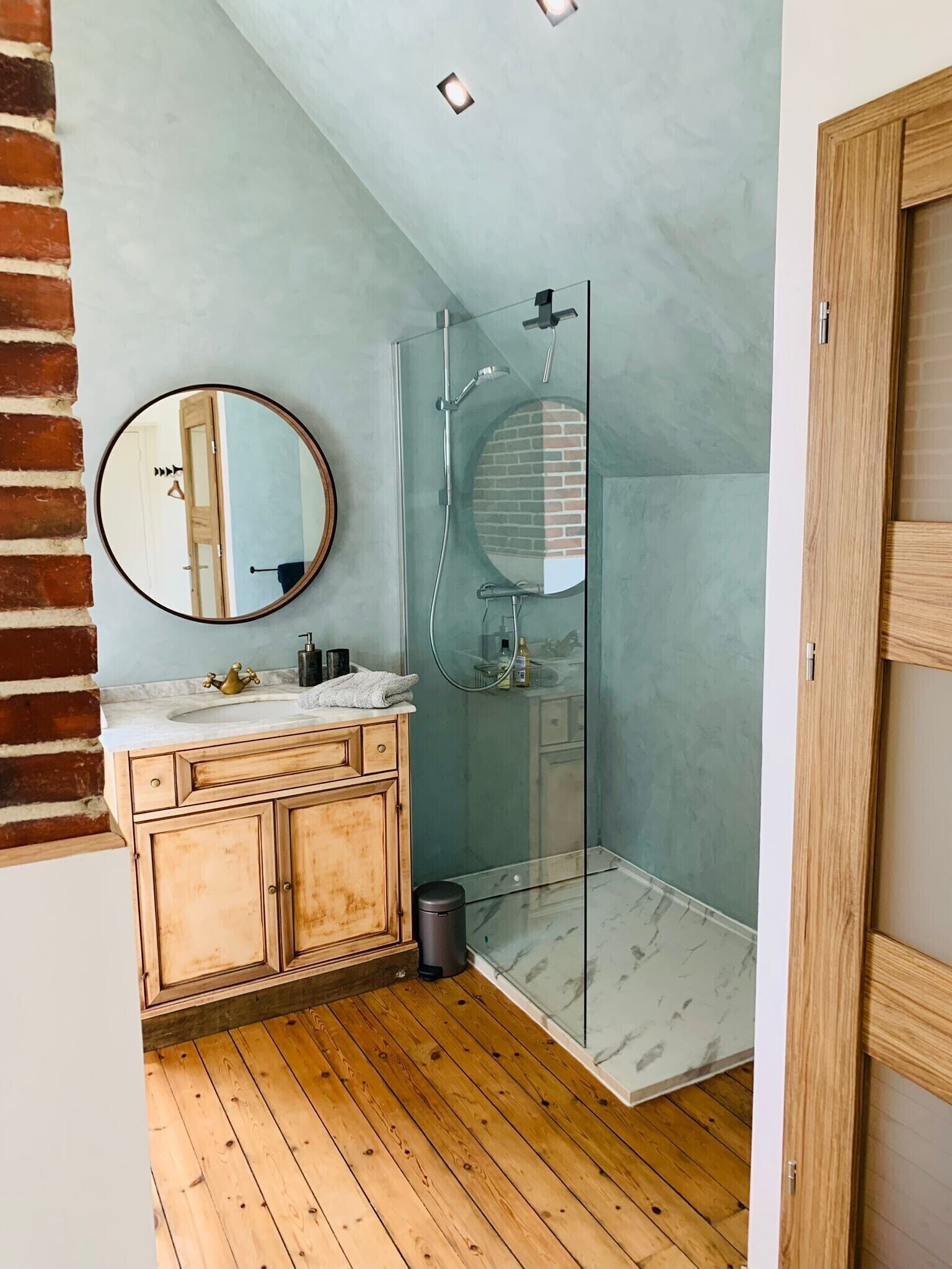 Une salle de bain avec un meuble-lavabo en bois, un miroir rond, une cabine de douche en verre et un plancher en bois.