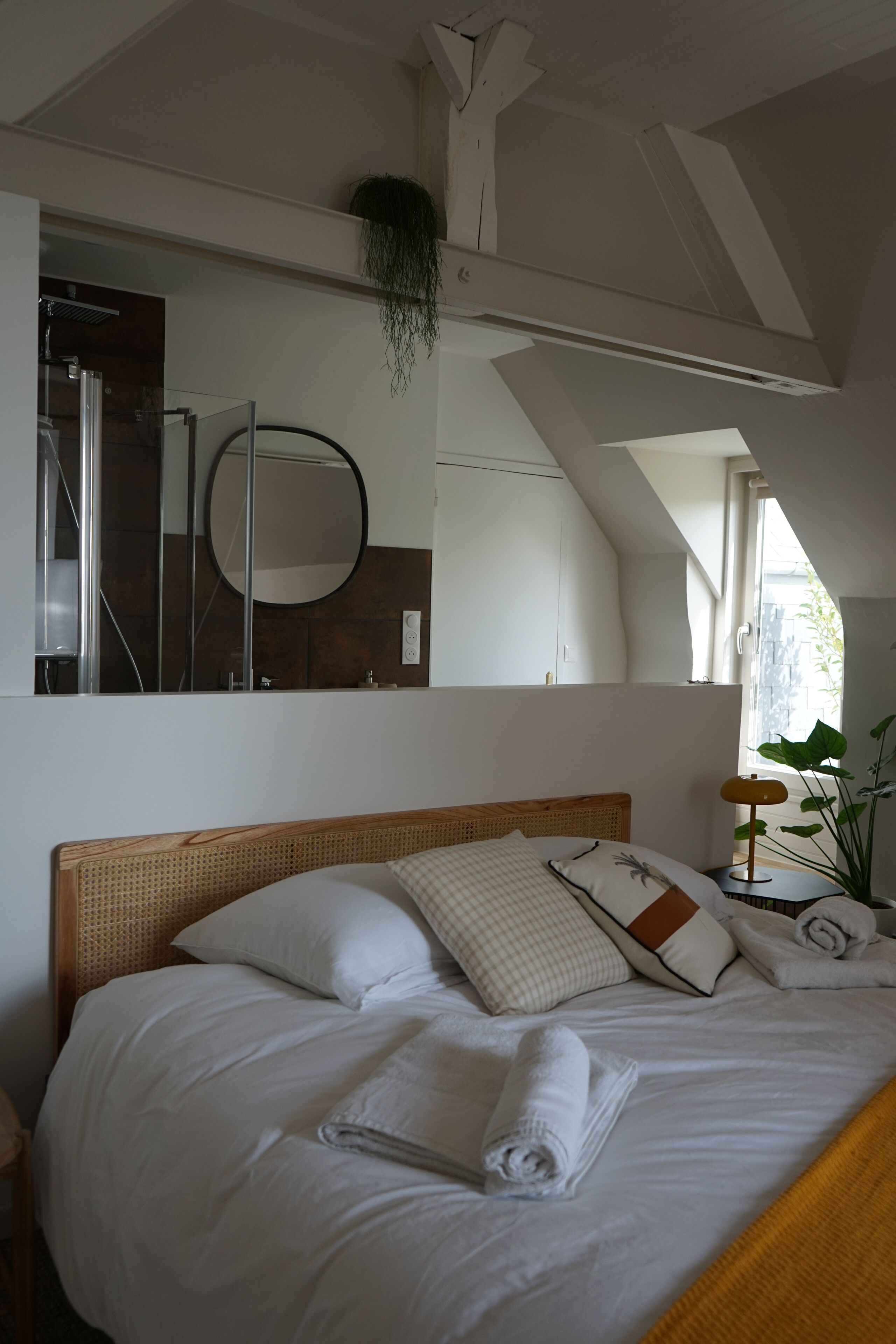 Ein ordentlich bezogenes Bett mit weißen Leinentüchern und einem zusammengerollten Handtuch, ein Nachttisch mit Lampe und ein großer Spiegel darüber.