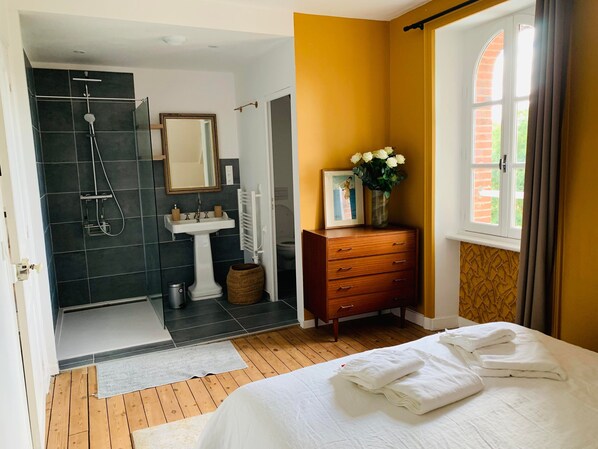 A hotel room with a wooden floor, a bed with white linens, a wooden dresser, a bathroom with a shower, a sink, and a mirror. - La Villégiature (Coutances)