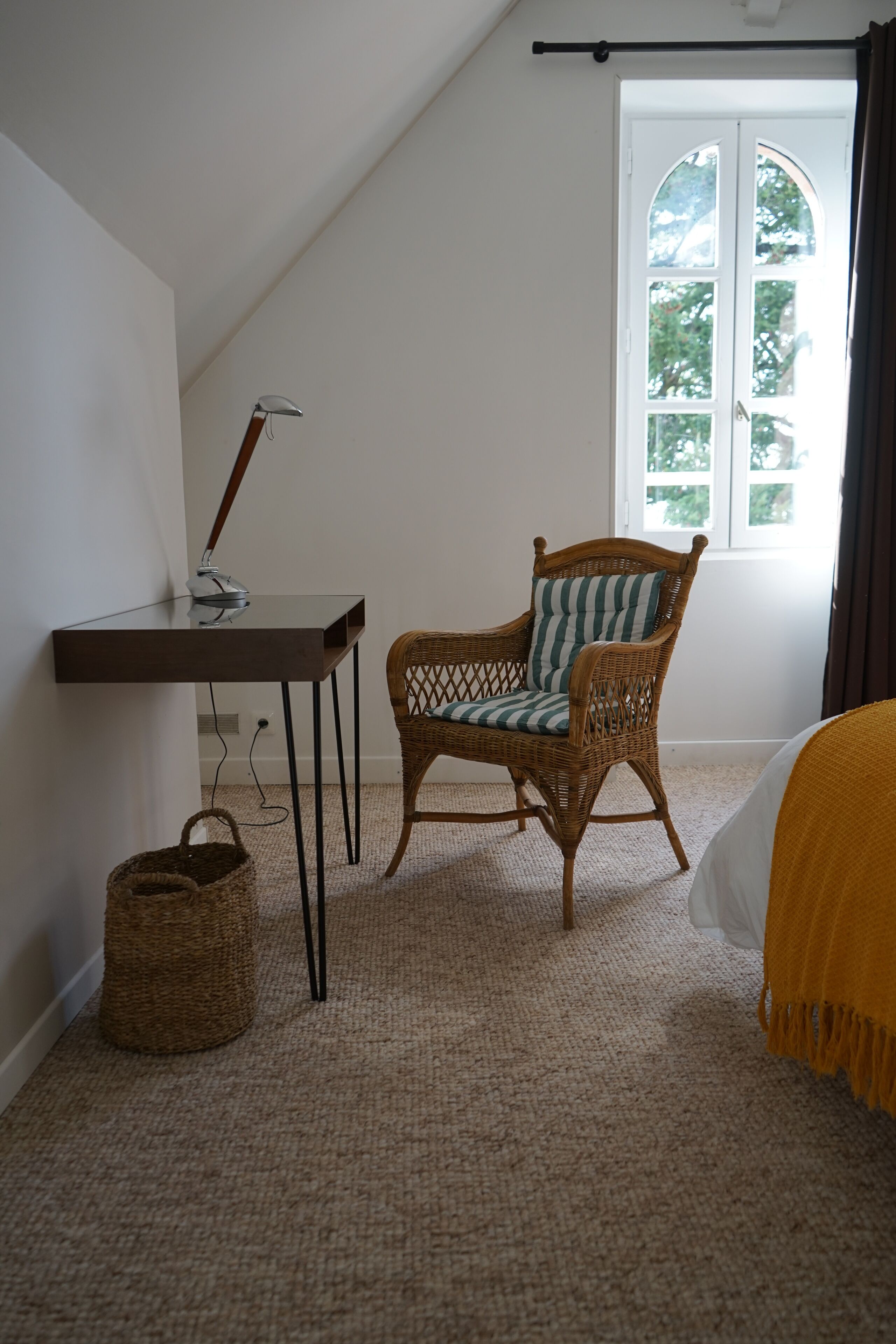 A bedroom with a sloped ceiling, a wicker chair, a small table with a lamp, a window with curtains, and a bed with a yellow blanket.