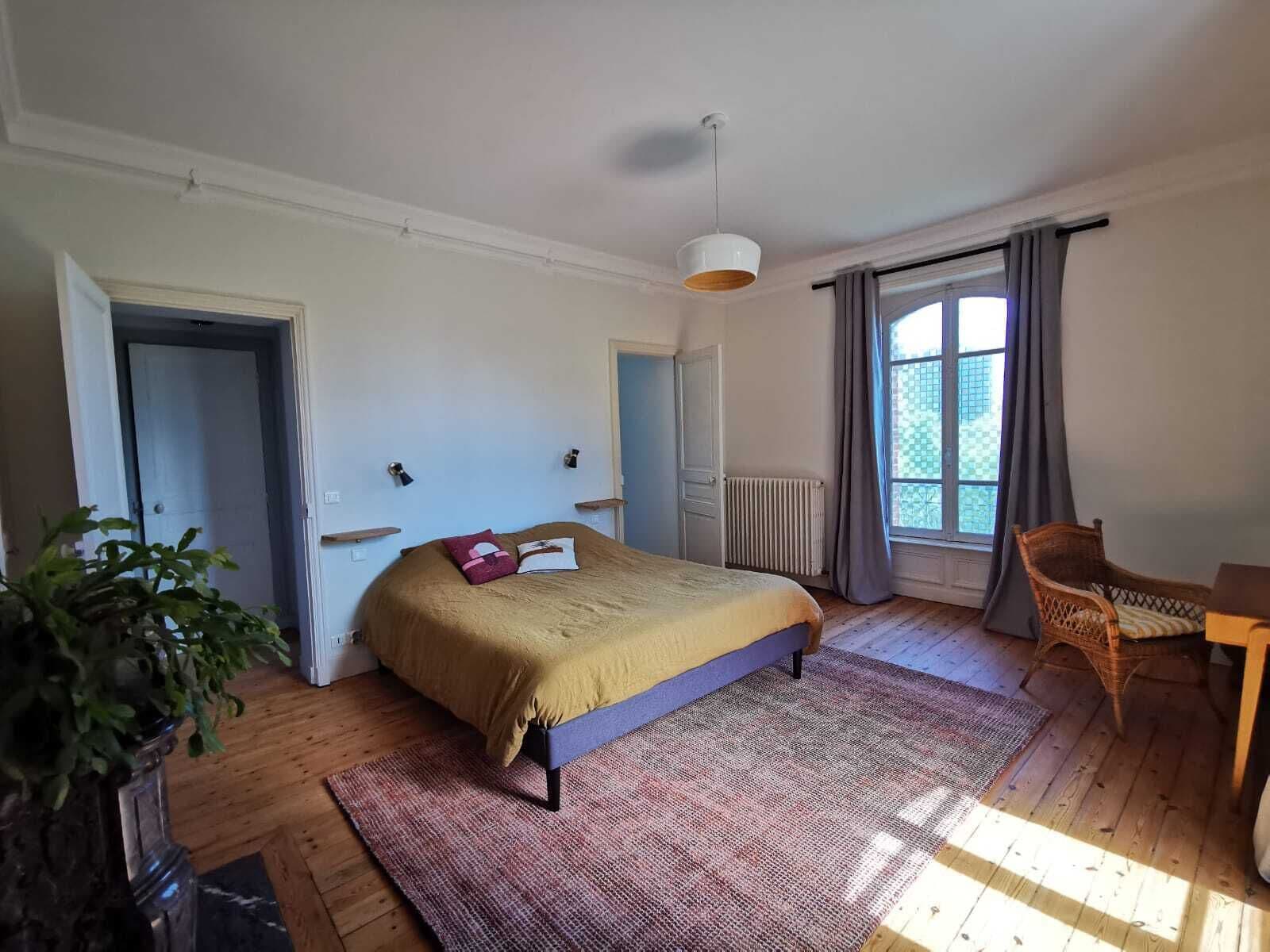 Een slaapkamer met een bed, een raam met gordijnen, een radiator, een stoel en een plant.