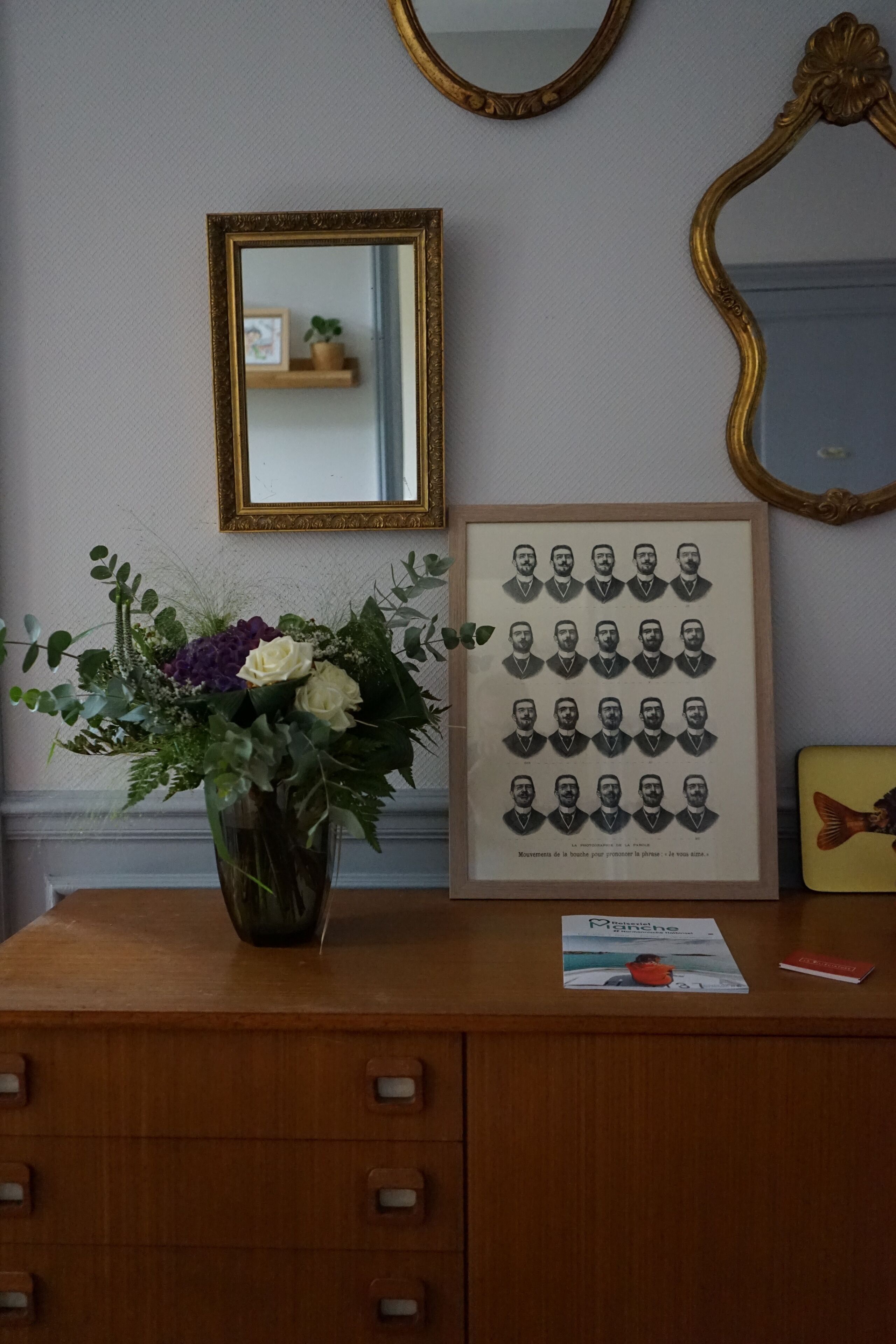 A wooden dresser with a framed photograph of individuals, a vase of flowers, and a mirror on the wall.