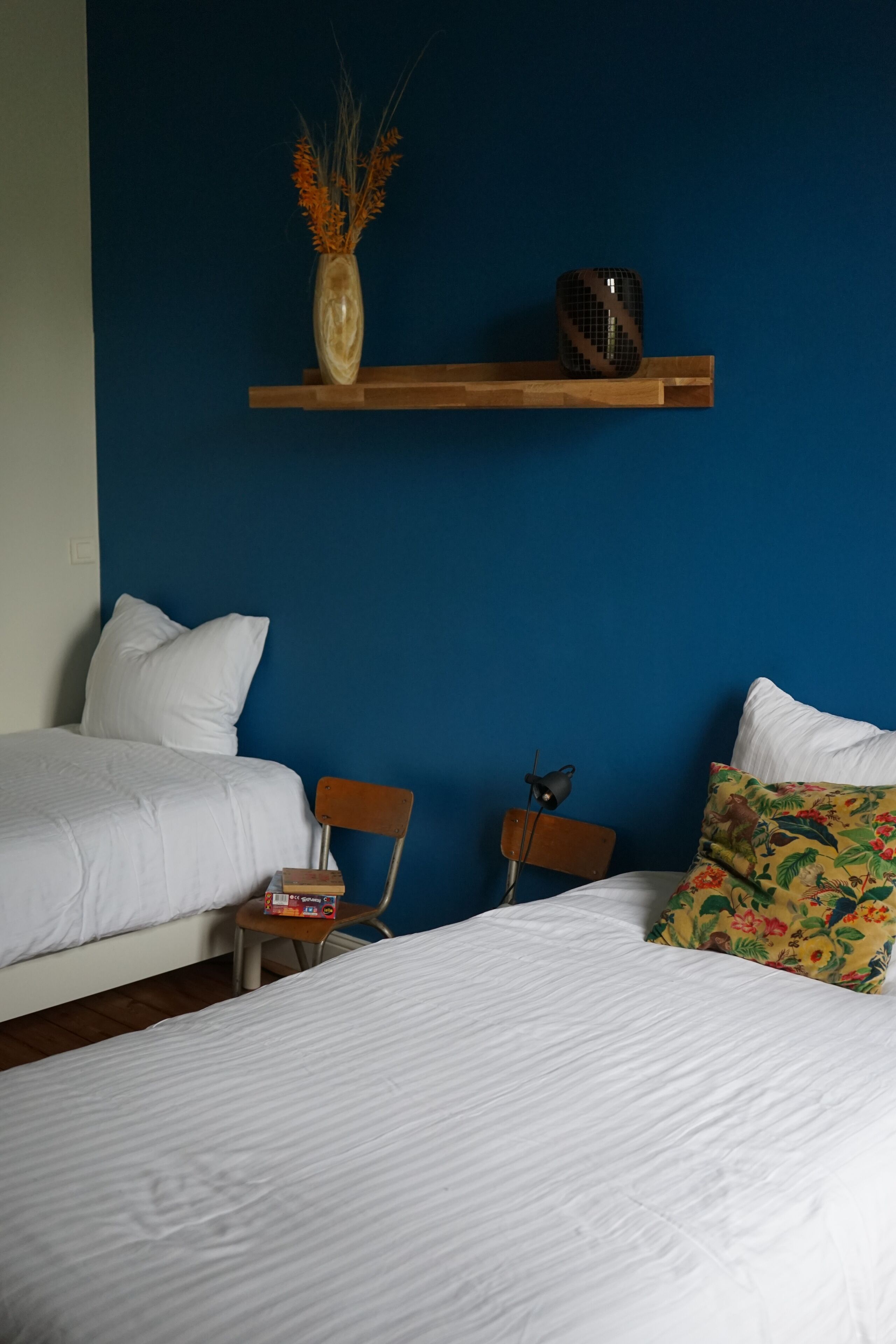 Une chambre d’hôtel avec deux lits, une chaise en bois, une petite table, un vase décoratif et une étagère sur laquelle se trouvent un ventilateur et une tasse.