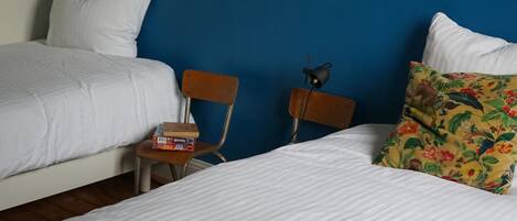 A hotel room with two beds, a wooden chair, a small table, a decorative vase, and a shelf with a fan and a mug.
