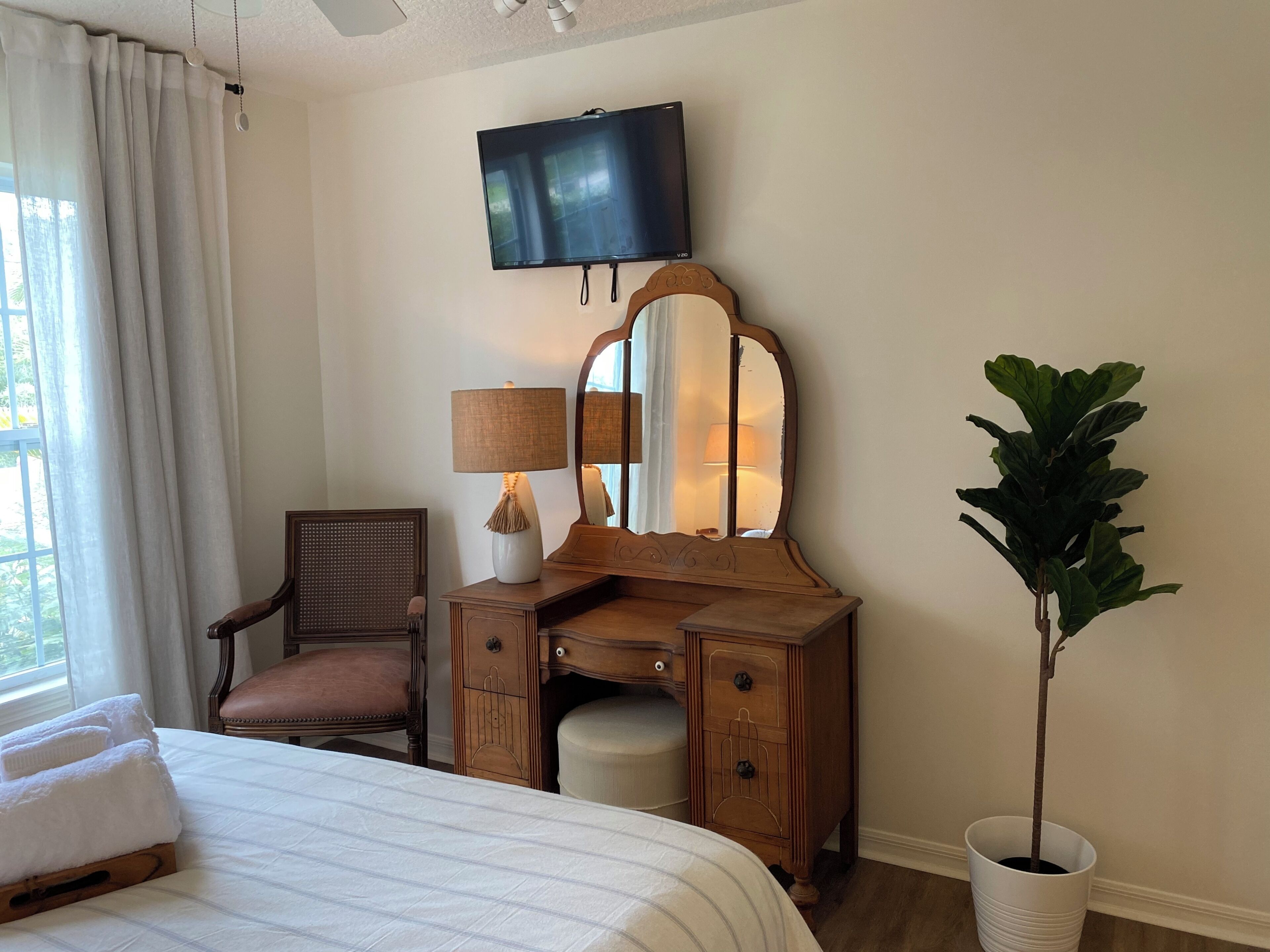 A hotel room with a bed, a wooden dresser with a mirror, a chair, a desk, a television, and a potted plant.