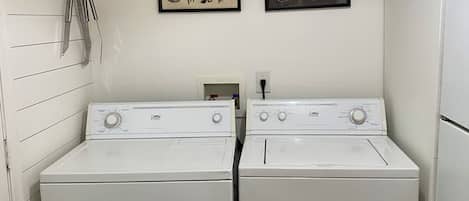 A laundry room with two white front-loading washers and dryers, a shelf with cleaning supplies, and two framed botanical prints on the wall.
