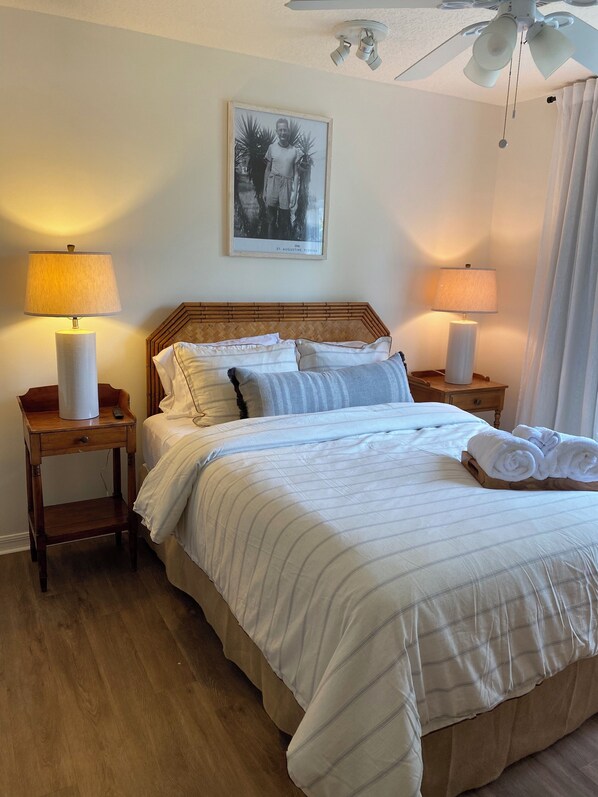 A neatly made bed with striped bedding, two bedside lamps, a framed photo on the wall, and a ceiling fan. - Old City Garden Retreat | Screened Porch • Walk to Historic St. Augustine (St. Augustine)